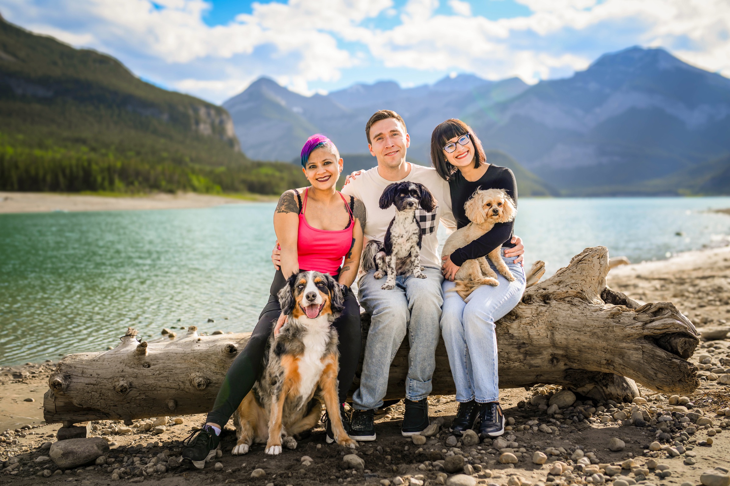 Three friends sit on a driftwood log by a turquoise mountain lake in the Canadian Rockies, holding their three dogs: an Australian Shepherd, a black and white spaniel mix and a fluffy apricot doodle