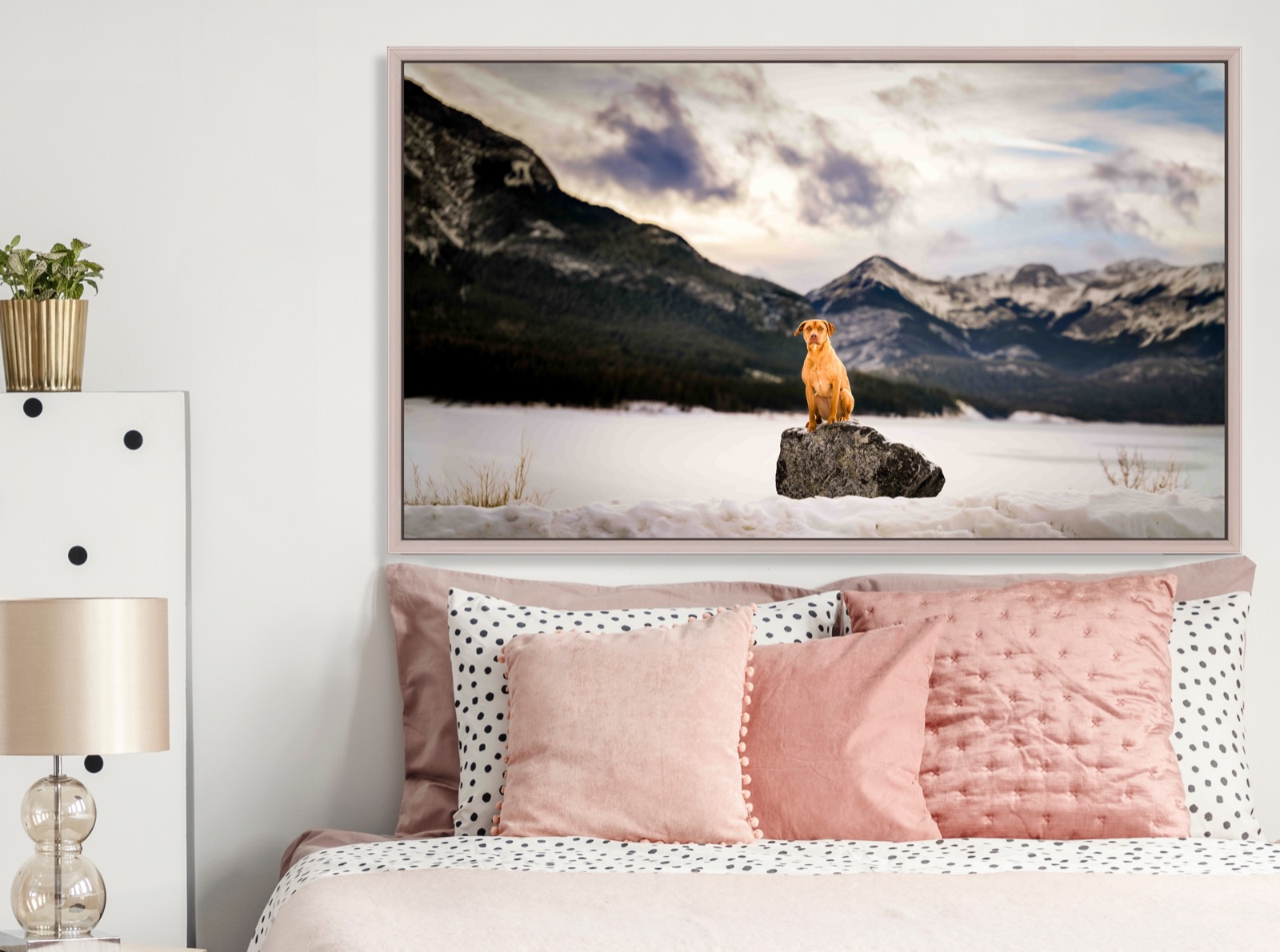 A large framed print of a red dog sitting on a rock in a snowy mountain valley, hanging above a bed with pink pillows